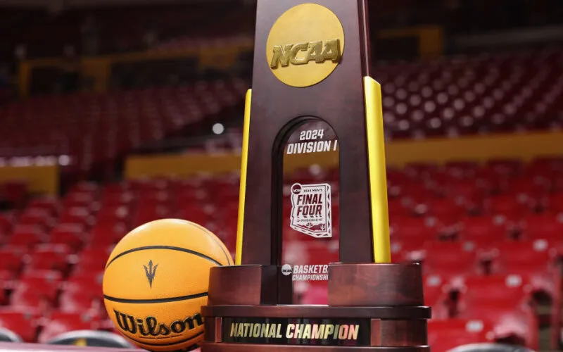 2024 NCAA Division I Men's Basketball Championship trophy displayed alongside a Wilson basketball on a basketball court, with empty red stadium seats in the background. The trophy features the NCAA logo, the label '2024 Division I Final Four,' and the words 'National Champion' on its base.