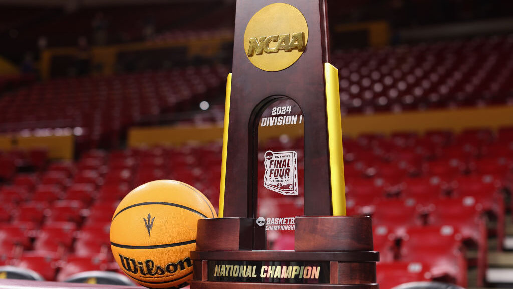 2024 NCAA Division I Men's Basketball Championship trophy displayed alongside a Wilson basketball on a basketball court, with empty red stadium seats in the background. The trophy features the NCAA logo, the label '2024 Division I Final Four,' and the words 'National Champion' on its base.