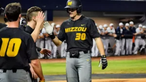 Mizzou Tigers baseball player wearing number 30 celebrates with teammates after scoring during a game, with the dugout visible in the background.