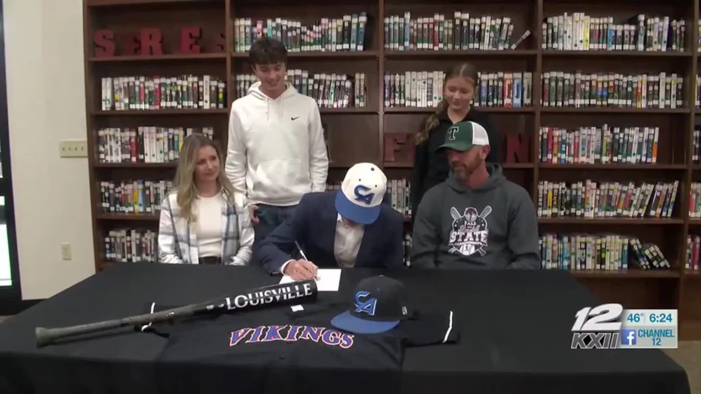 A Tushka High School baseball player signs a college commitment letter surrounded by family and coaches in a library setting. The player, wearing a Carl Albert State College cap, is seated at a table decorated with a Louisville Slugger bat, a Vikings jersey, and team caps.