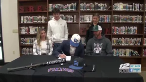 A Tushka High School baseball player signs a college commitment letter surrounded by family and coaches in a library setting. The player, wearing a Carl Albert State College cap, is seated at a table decorated with a Louisville Slugger bat, a Vikings jersey, and team caps.